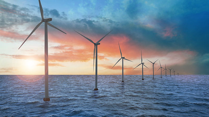 A row of offshore wind turbines, stretching into the distance across the ocean under a partly cloudy sky with hues of blue, orange, and pink.