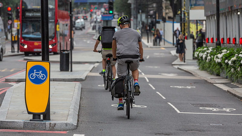Two cyclists ride on a segregated bike lane in London, marked with white bicycle symbols and separated from the main road by a curb. A yellow sign with a blue bicycle symbol is visible on the left. In the background, pedestrians walk near buildings and a red double-decker bus.