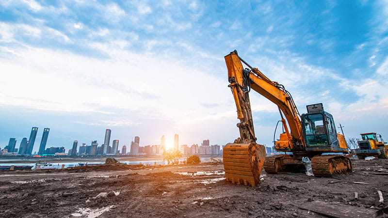 A construction site near a body of water with two excavators, one orange in the foreground and another in the background. A city skyline with tall buildings is visible under a partly cloudy sky during sunset or sunrise.