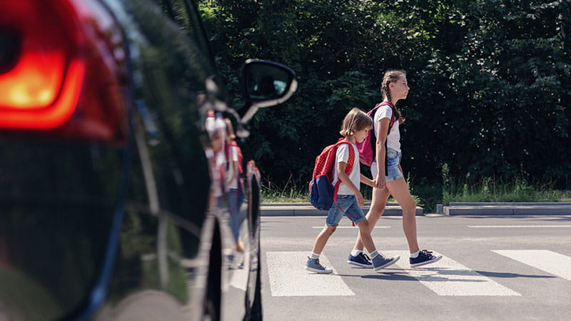 Two children, one older and one younger, cross a street at a zebra crossing. A car with its brake lights on is in the left foreground, and the background features greenery and trees.